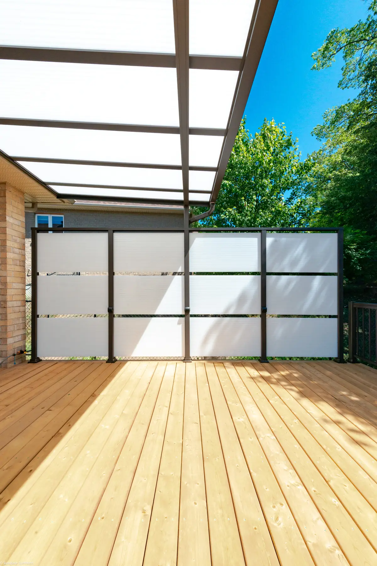 A modern Privacy Wall with frosted panels and a dark metal frame on a wooden deck, partially shaded by a clear Patio Cover.