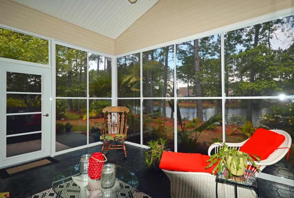 A cozy enclosed patio featuring wicker and wooden furniture, panoramic lake views, and lush greenery outside.