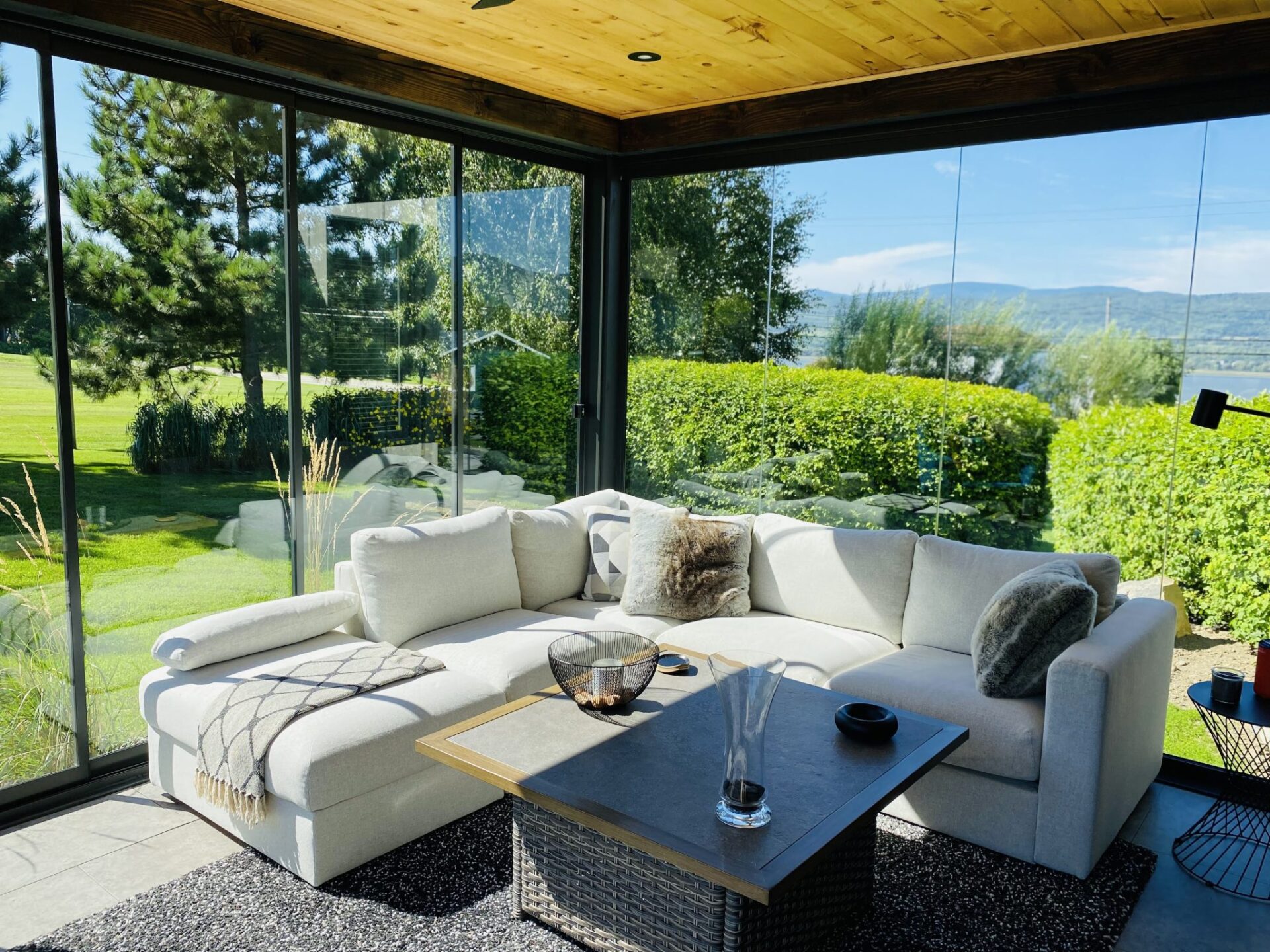 The Horizon Plus Sunroom by Novasun showcases a sleek black frame, large glass walls, and elegant wooden furniture, nestled in a lush, vibrant backyard garden.