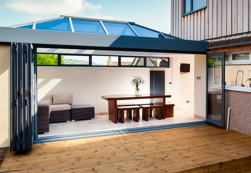 Patio with glass roof and dining space featuring WeatherMaster™ Plus Bi-Fold Doors for effortless access.